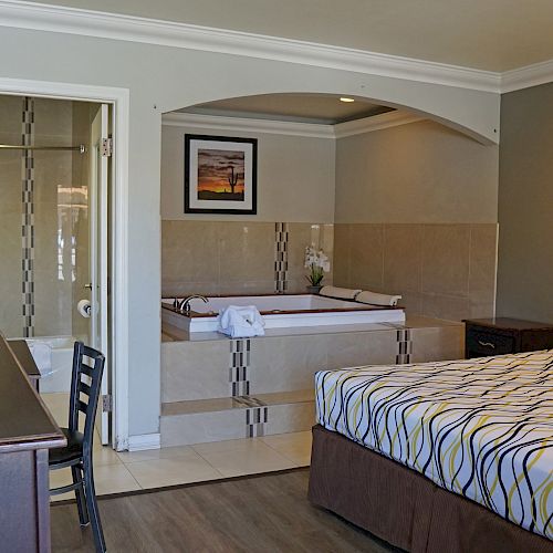 A hotel room with a bed, desk, chair, TV, and an en-suite bathroom visible, featuring a bathtub under an archway.