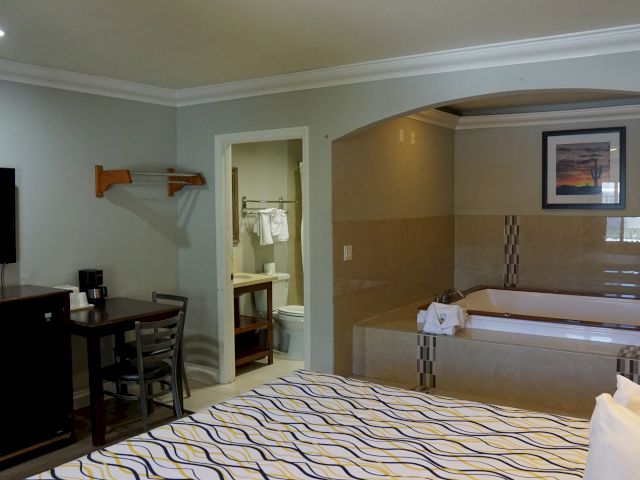 A wide view of a hotel room with a bed, table, and bathroom with a large tub.