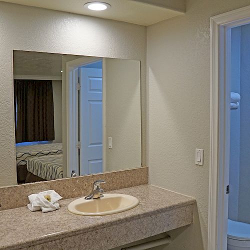 A bathroom with a sink, mirror, and countertop featuring towels and toiletries. A shower and toilet are visible in an adjacent room.