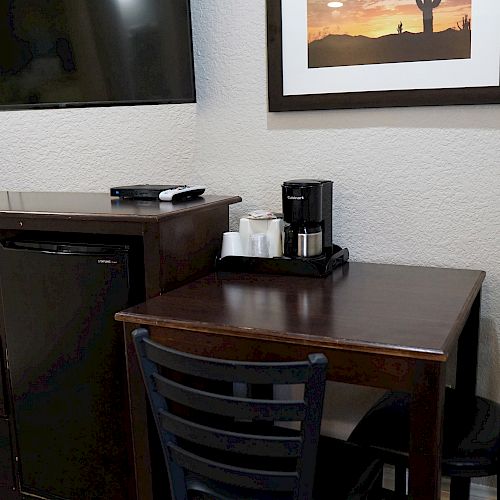 The image shows a small dining area with a table, two chairs, a coffee maker, a microwave, a mini-fridge, and a wall-mounted TV.