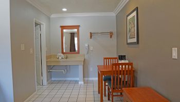 The image shows a hotel room with a sink area, mirror, table, chairs, framed artwork, and a small set of drawers. The room has gray walls and tiled floor.