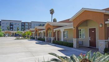 The image shows a row of single-story units with arched doorways and an empty parking lot in front, with an apartment building in the background.