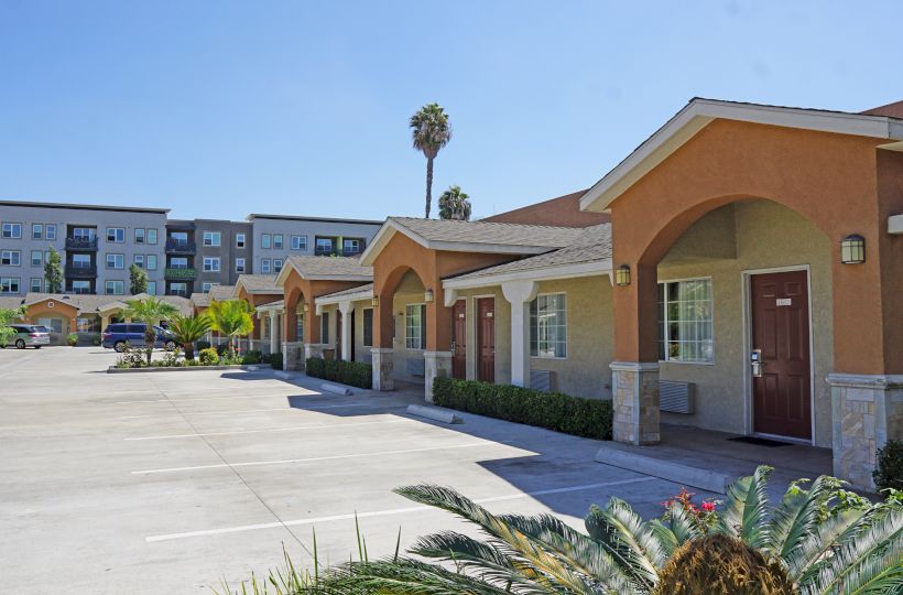 The image shows a row of single-story units with arched doorways and an empty parking lot in front, with an apartment building in the background.