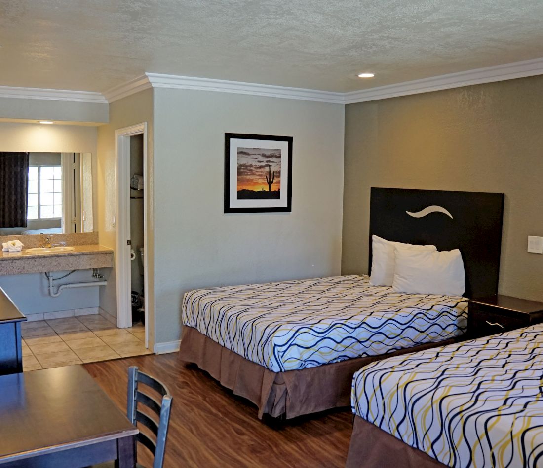 A hotel room with two beds, a nightstand, a table with a chair, and a bathroom in the background.
