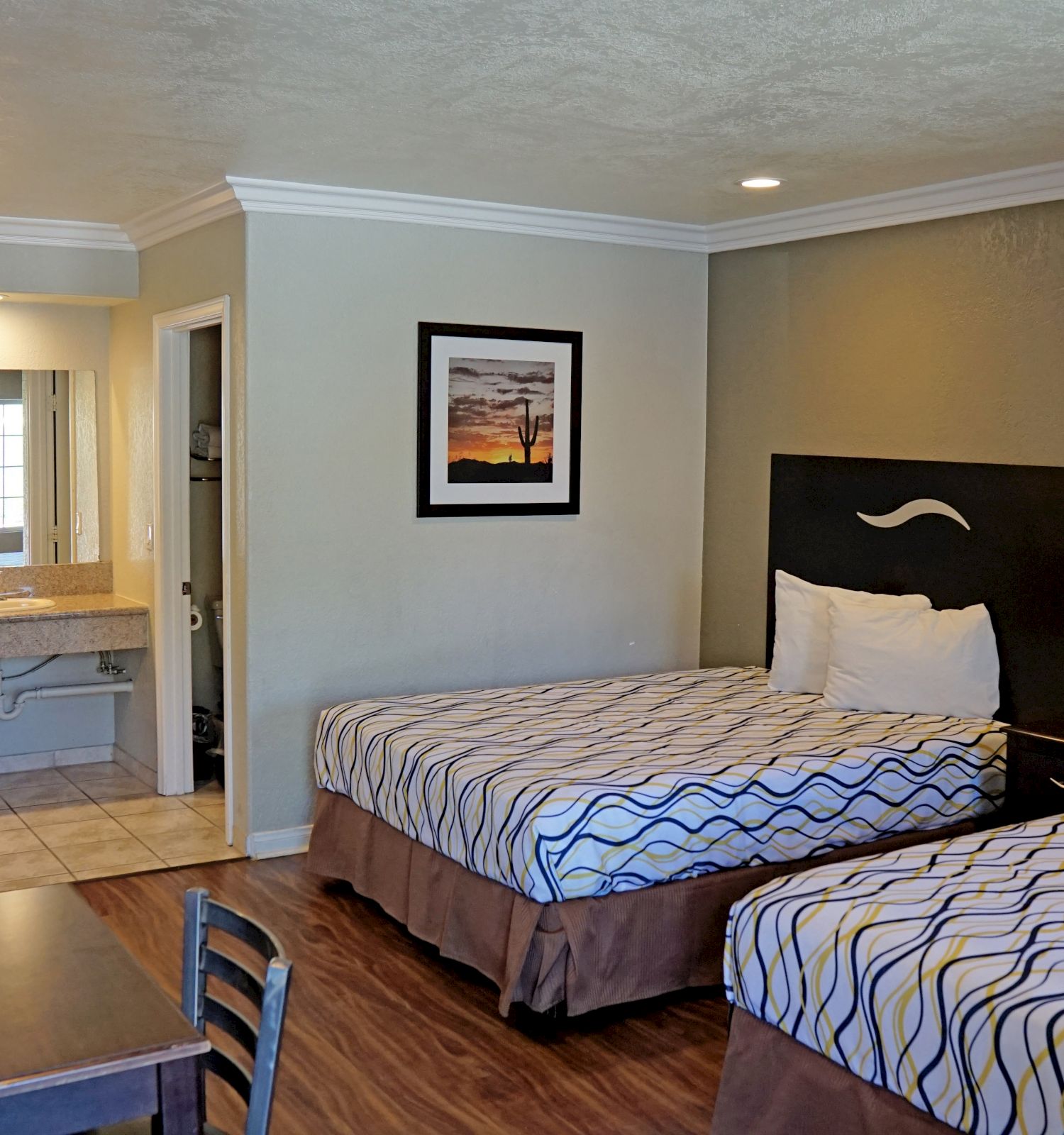 A hotel room with two beds, a desk, a TV, and a vanity area with a sink leading to a bathroom. The room has minimal decor and wood flooring.