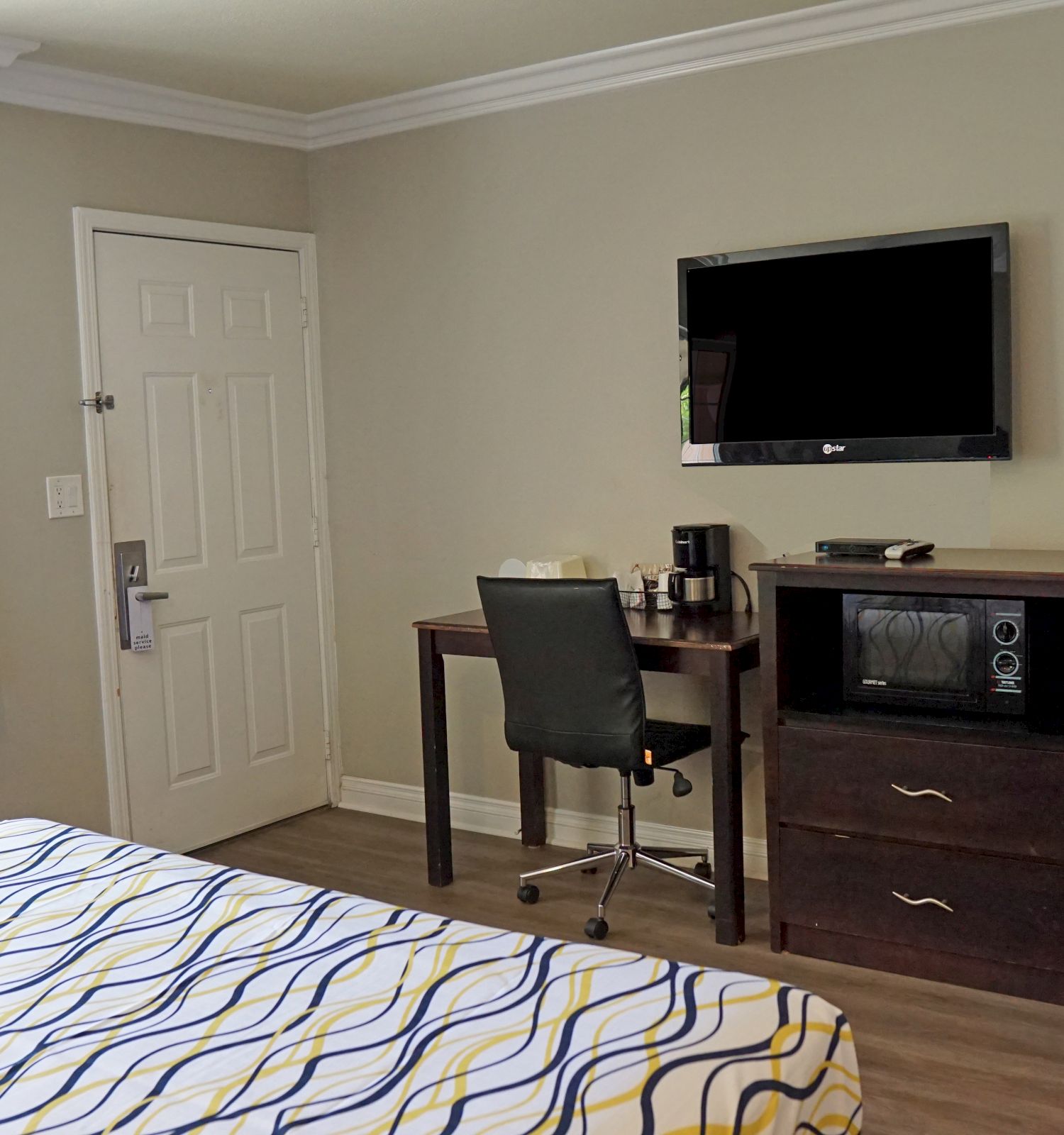 A hotel room with a bed, desk, TV, microwave, mini-fridge, and coffee maker. The room has a window, a door, and an air conditioning unit.