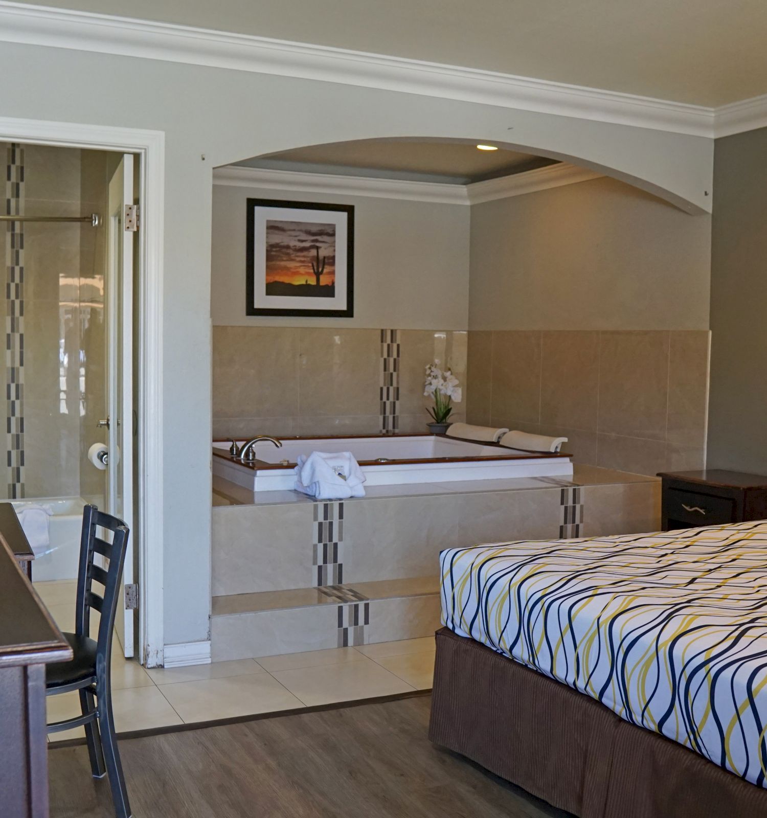 The image shows a hotel room with a large bed, a desk with a chair, a TV, and a jacuzzi in an alcove next to a bathroom with a shower.